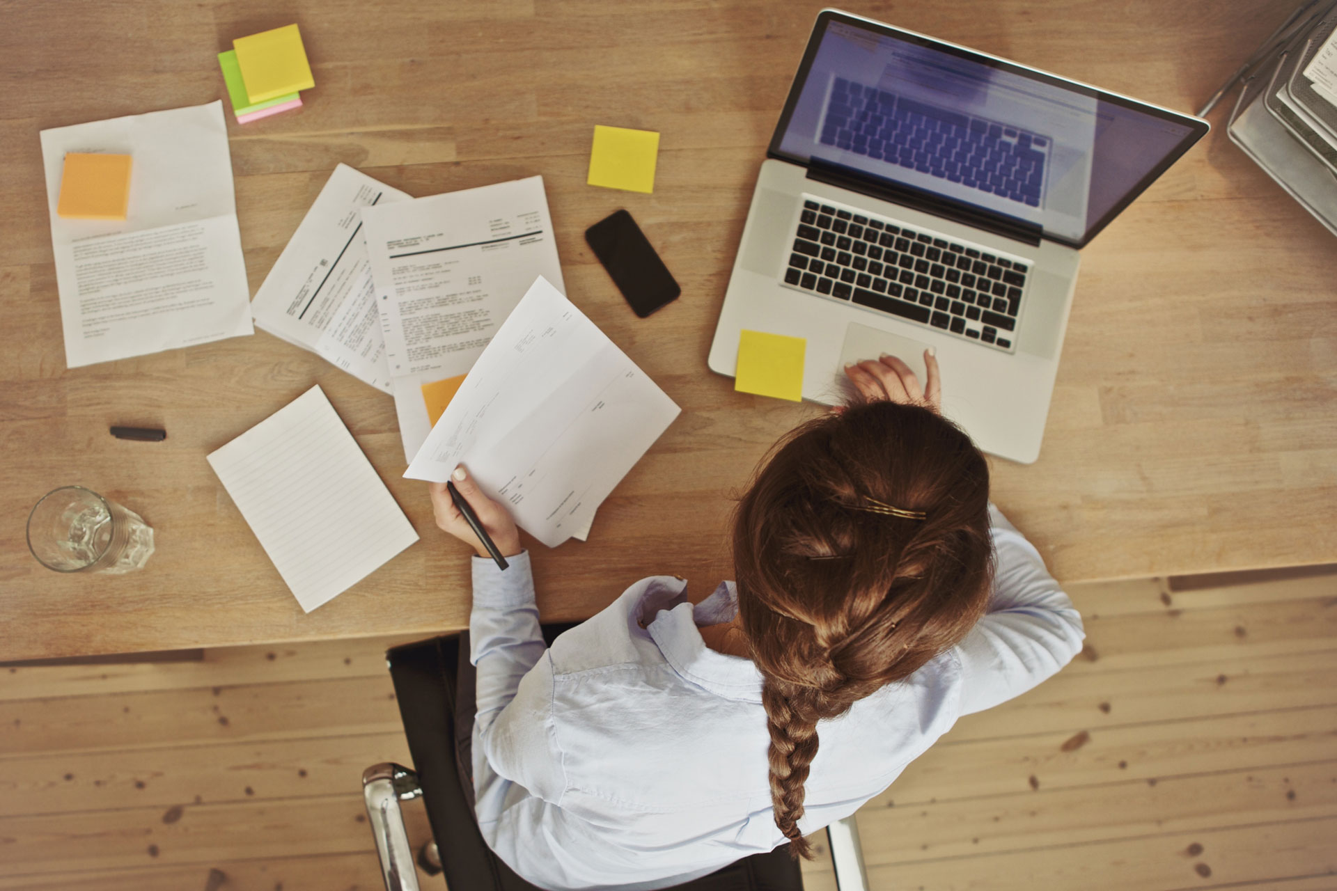 Person with braided hair works on laptop and reviews papers at wooden desk with notes and phone.