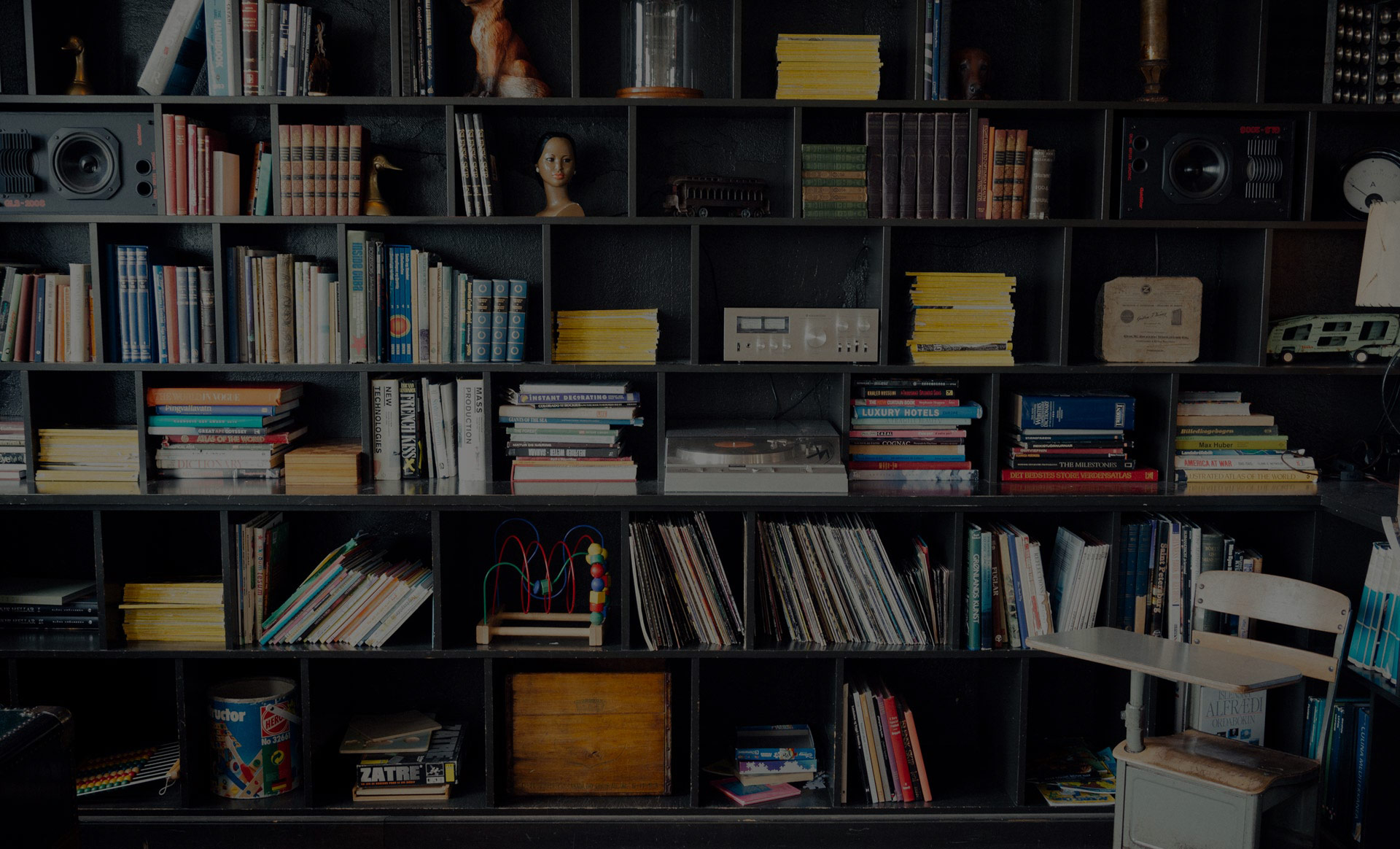 Bookshelf with books, magazines, vinyl records, vintage record player, toy bead maze, and a wooden chair with attached desk.