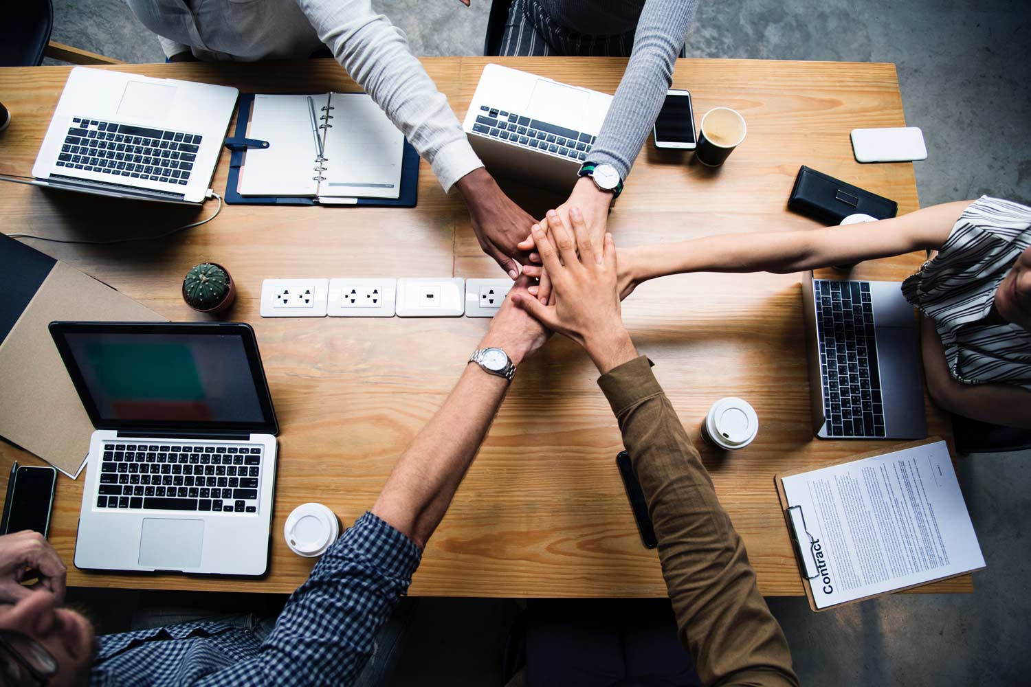 Four people join hands over a wooden table with laptops, documents, phones, and coffee cups.