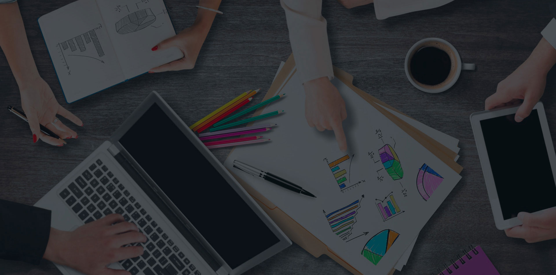 Workspace with laptop, tablet, charts, colored pencils, coffee cup, and hands interacting.